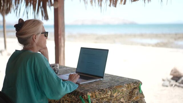 A mature woman sits under a palapa using a computer while enjoying the tropical beach in Baja California, Mexico, during the day.