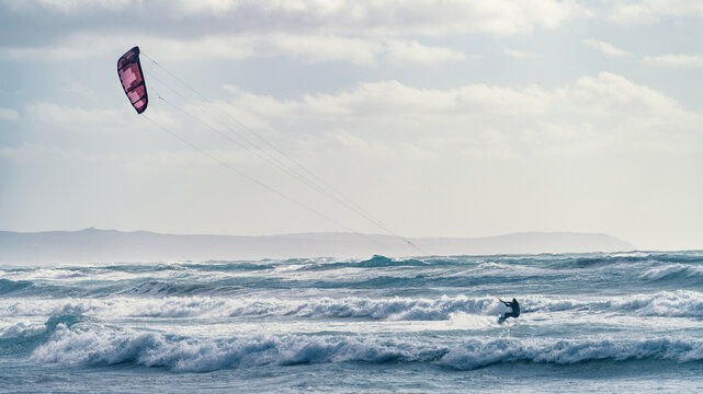 View of a kitesurfer riding over choppy waves with a pink kite under a cloudy sky in Sardinia, Italy.