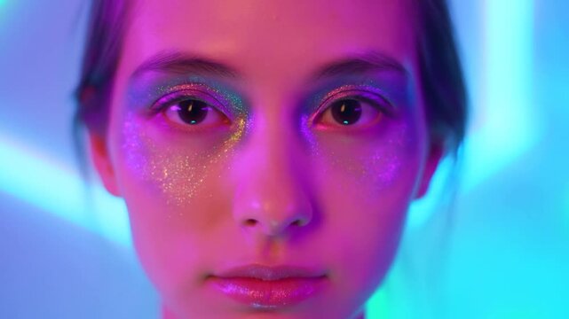 Close up slow motion tracking shot of young woman blinking with glitter makeup and colorful neon lighting against soft studio backdrop frontal portrait