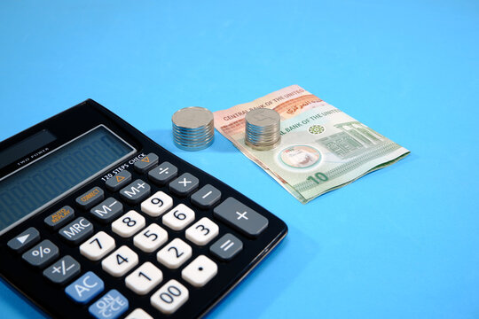 A desktop calculator placed next to a ten dirham banknote and stacked silver coins isolated on a blue background representing financial accounting, business investment, savings management, budget plan