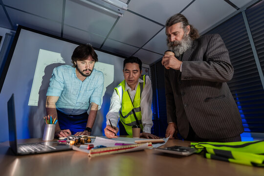 Team of professionals reviewing plans together in indoor workspace with equipmen. Architect and engineering team having a late-night meeting and presentation for a real estate project in the office.