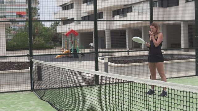 Young woman playing padel on an outdoor court in a residential complex in Spain. Female athlete training near the net during a practice session. Active lifestyle, fitness and racket sport concept