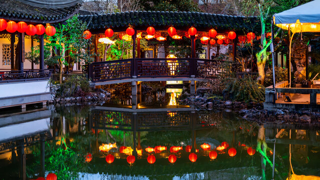 View of Lan Su Chinese Garden with red lanterns hanging from a traditional bridge reflected in a pond at dusk Portland, Oregon, United States.