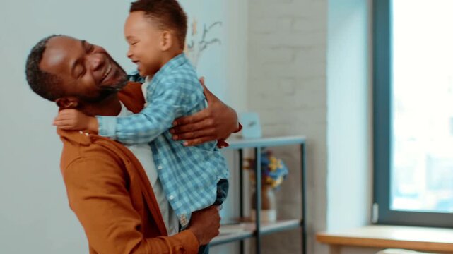A man with dark skin holds a smiling boy in a blue shirt. They are indoors, enjoying a playful moment together. The room has natural light and a cozy atmosphere, promoting connection.