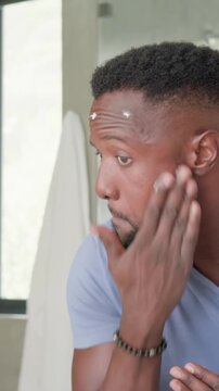 Vertical video: Seeing cream dots, man rubbing cream at bathroom mirror in blue tee for skincare