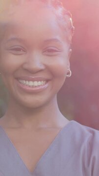 Vertical video: Posing woman widening grin as sun brightens lens flare in park, gray V-neck, hoops
