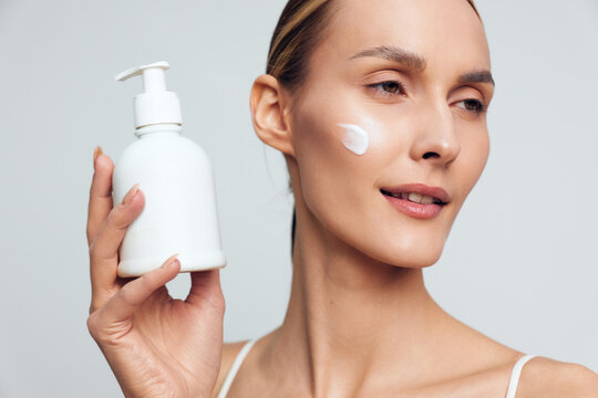 Woman with face cream, skincare self care, holding pump bottle, close up portrait, white cream on cheek, facial wellness, soft studio lighting, clean beauty, calm smile
