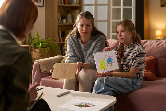 Caucasian mother and girl showing drawings while meeting therapist, discussing child emotions and family support during counseling session. Useful for mental health, parenting, therapy marketing