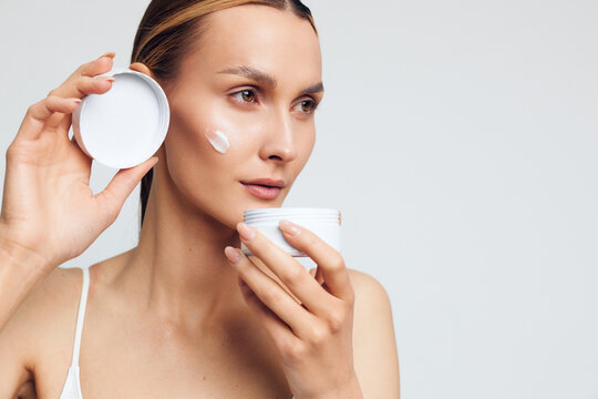 Woman skincare beauty face cream cotton pad self care hygiene, applying moisturizer to cheeks, gentle touch, focused gaze, soft studio lighting, minimal white background fresh skin