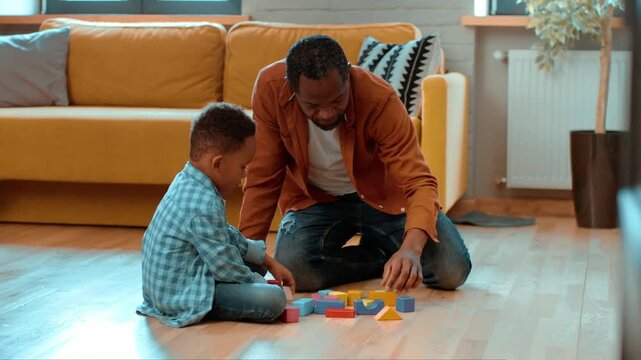 A father interacts with his son as they play with colorful building blocks on the floor of a cozy living room. They focus on building structures while enjoying their time together.