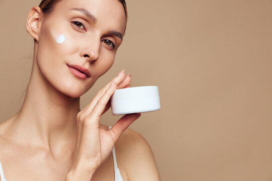 Close up of a woman applying face cream for beauty hydration, beige background, calm smile, skincare self care concept, wellness, gentle moisturizer, studio lighting