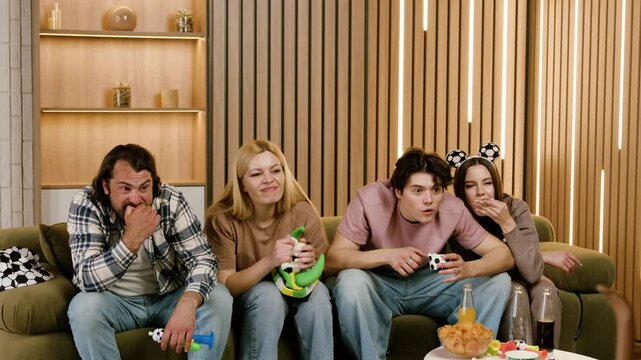 Group of young adults watching sports on a couch, displaying tension and excitement as they react to the game, snacks and drinks on the table in a cozy living room setting
