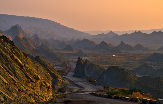 View of the Makran Coastal Highway winding through jagged rock formations and rugged desert mountains under a warm golden sunset sky in Makran, Balochistan, Pakistan.
