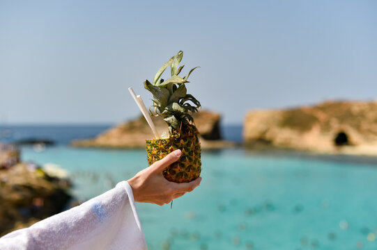 A person holds a cocktail served inside a hollowed-out pineapple with two straws. The background shows a blurred turquoise sea lagoon and rocky cliffs.
