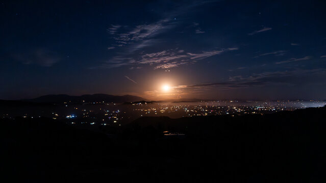 View of the glowing city lights of Fallbrook nestled in a valley under a bright moon and a starry night sky with wispy clouds in Fallbrook, California, United States.