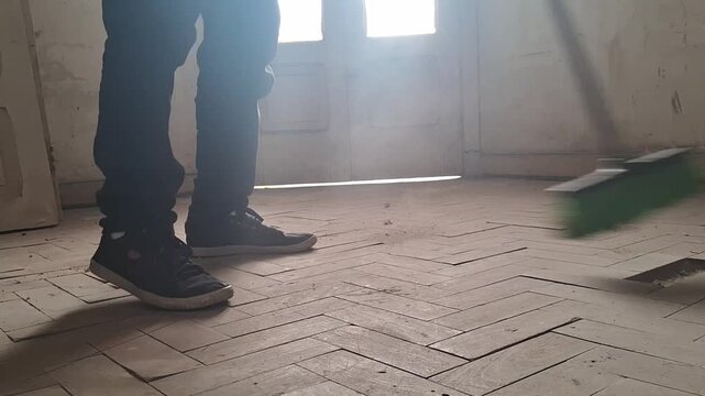 Individual sweeping a dusty, old herringbone parquet floor with a green broom, renovation video.