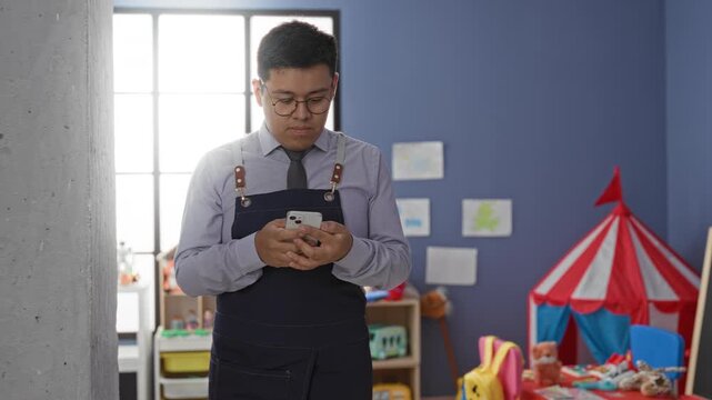 Man teacher texting on smartphone with hands visible, wearing apron and tie in a kindergarten building; quiet concentration.