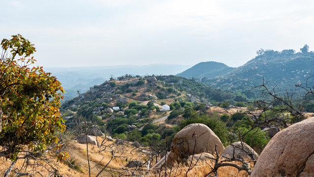 View of the rocky hills of Splitrock Baby Oaks with large boulders, green vegetation, and a white dome structure under a hazy sky in Fallbrook, California, United States.