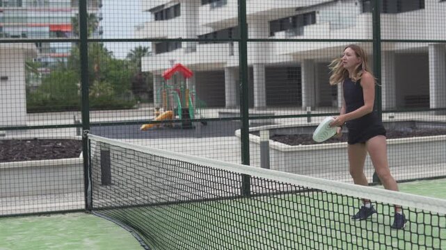 Young woman playing padel and hitting a volley near the net on an outdoor court in Spain. Focused action moment during a training session in a residential complex