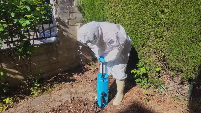 Female gardener in white protective suit operates manual hand pump to pressurize blue pesticide backpack sprayer in garden.