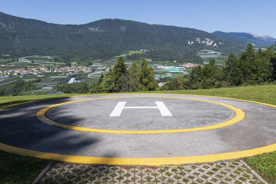 View of a concrete helipad with a white letter H and yellow border overlooking a lush valley with a small village and mountains under a clear blue sky in Italy.