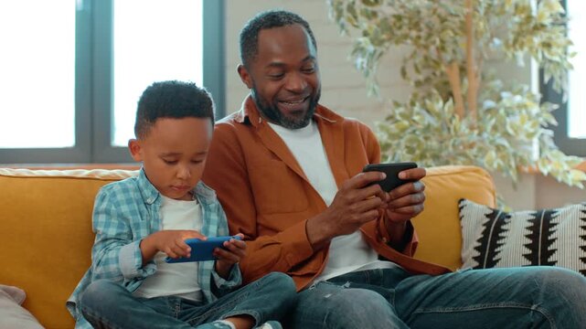 A Black man and a young boy sit side by side on a couch. They both are focused on their smartphones while relaxing in a bright and cozy living room. The atmosphere feels warm and inviting.