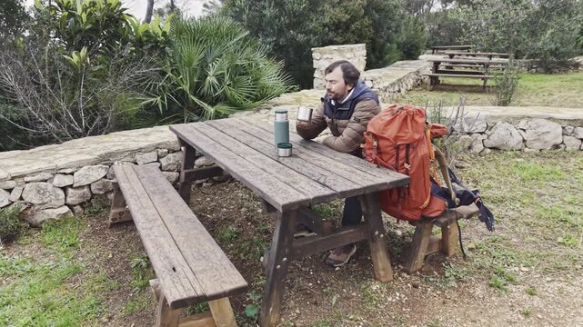 Man resting at picnic table drinking from mug during hiking trip.