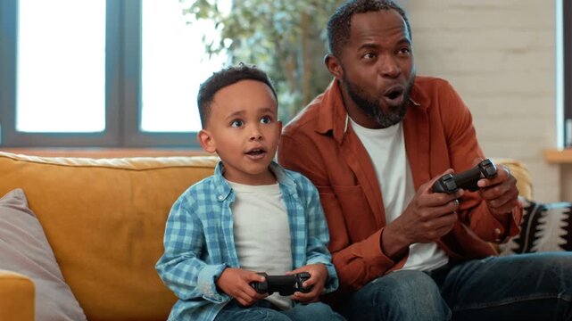 A man with dark skin and a boy are engaged in playing video games. They both hold controllers and show excitement while enjoying their time together at home.