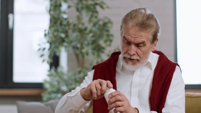 A senior man with gray hair and a beard looks closely at a bottle of supplements while seated in his living room. The setting features plants and natural light coming through a window.