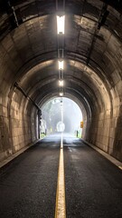Obraz premium Long tunnel with a bright exit. The walls are concrete, and the road has a yellow dividing line. Light fixtures are ceiling mounted