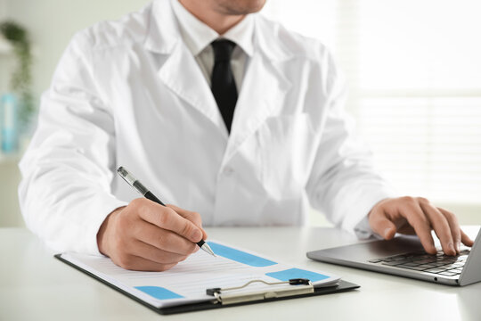 Doctor in medical coat working with clipboard and laptop at white table in clinic, closeup