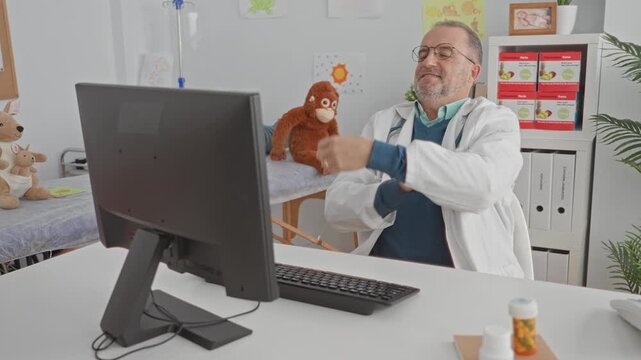 Man doctor wearing stethoscope leans back with arms crossed at pediatric clinic building while seated at computer desk; contentment.
