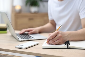 Young student studying with laptop and writing in notebook at wooden table indoors, closeup