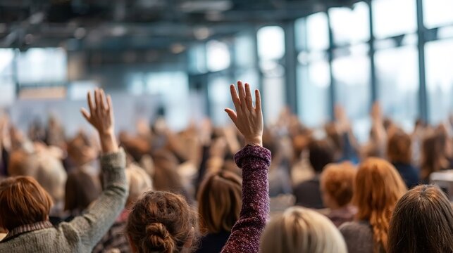 People raising hands from their seats in a blurred conference hall, actively participating in a question and answer session during a corporate event or educational presentation