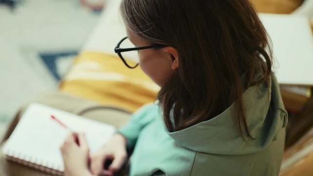 Smart Little Girl in Big Glasses Writing in Notebook while Sitting on Couch with Stack of Books &mdash; Cinematic 4K 10-bit