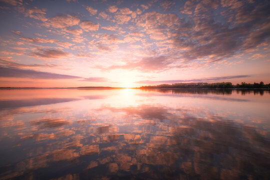 View of a sunset over a calm lake with pink clouds reflecting on the water surface and a distant tree-lined shore in Piaski, Warmian-Masurian Voiv, Poland.