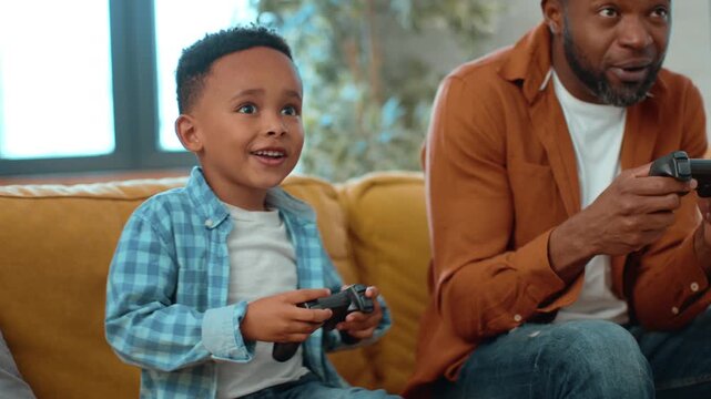 A man with dark skin and a boy are playing video games in a bright living room. The boy smiles as they both hold game controllers. They share a fun moment on a weekend at home.