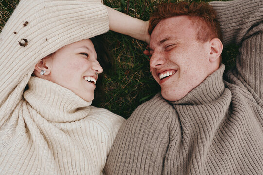 Two people lie on grass wearing neutral sweaters and smile at each other, creating a cozy lifestyle moment outdoors, with warm light highlighting connection, comfort, and happiness in a candid,
