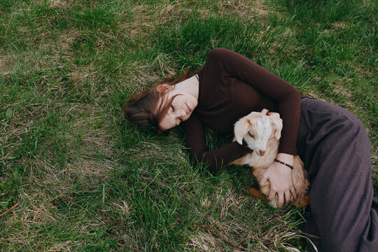 A woman rests on grassy ground, cuddling a small goat kid in a peaceful outdoor setting. The moment captures gentle companionship, rural lifestyle, and tender bonding between human and animal.