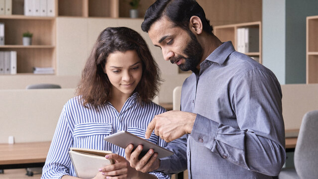 Diverse friendly coworkers colleagues indian male manager and latin female employee talking in office space using tablet device. Effective management, teaching and mentoring new employee concept.