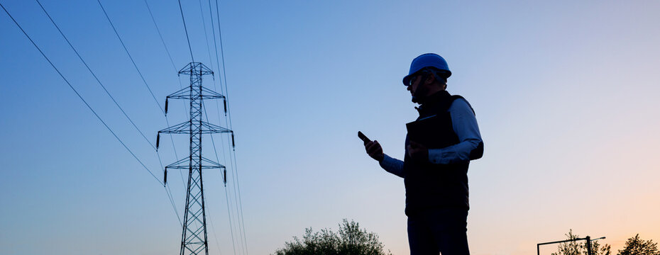 Energy worker in blue hard hat and gilet inspecting power lines infrastructure. Banner - silhouette of a male engineer against a background of high-voltage power transmission line poles. 
