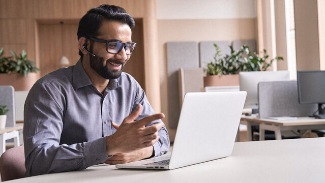 Happy indian business man remote teacher, customer support manager wearing headset talking at virtual meeting consulting client on video call giving distance learning class at home office call center.