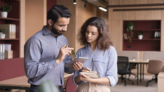 Indian manager holding digital tablet having discussion with latin employee meeting in office lobby. Two diverse professionals talking using pad discussing project strategy, checking corporate plan.