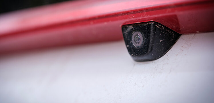 Extreme close up capturing a small exterior backup reversing camera integrated discreetly beneath a red decorative trim element on a modern white vehicle trunk