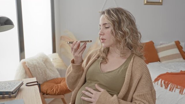 Pregnant woman holding smartphone to mouth in bedroom, hand on belly while speaking into device; anticipation connection preparation.