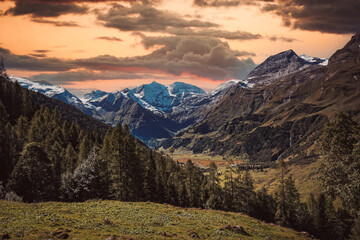 Fototapeta premium Peaceful picturesque mesmerizing atmospheric landscape with high Snow-capped mountains, Großglockner alpine serpentine pass road, and meadow grasslands pasture in sunset