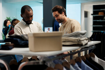 Fototapeta premium Customer and assistant discussing outfit while browsing clothing rack. Shopping mall boutique shopper receiving guidance from employee while examining garment before purchase