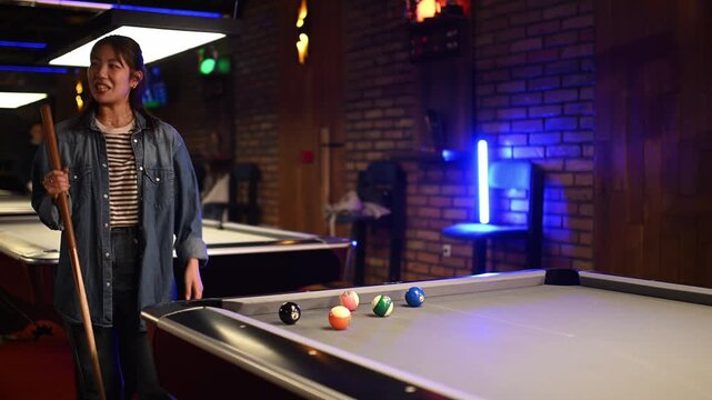Young asian woman hitting pool balls with a cue stick, enjoying a game of billiards in a modern bar with neon lights