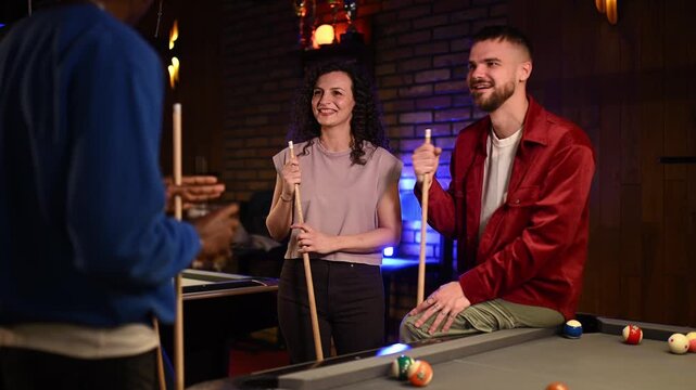 Young multicultural friends enjoying a game of billiards. Men and women talking, laughing, and holding cues around a pool table at night
