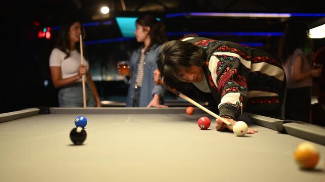 Focused young man shooting the cue ball while playing billiards with friends in a bar at night